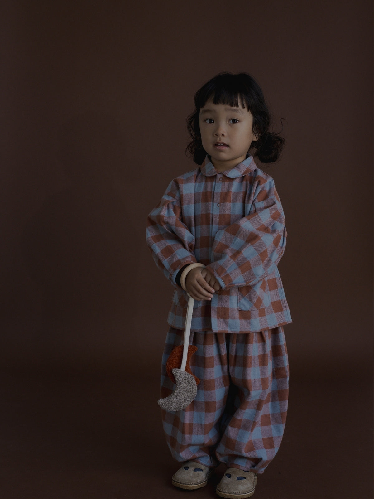 A young child stands against a dark brown studio background wearing a matching plaid set in dusty pink and light blue. The outfit consists of a long-sleeve button-up top and loose pants, and the child is holding a small grey crescent moon toy.