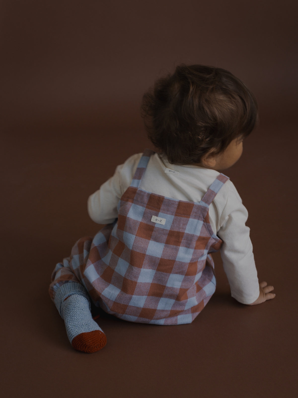 A baby sitting on a brown studio floor, viewed from behind, wearing a white long-sleeve top and rust-orange and light blue gingham check overalls.