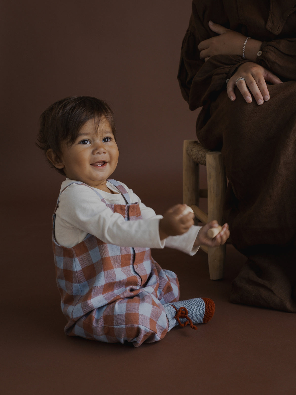 A baby wearing a white long-sleeve shirt and a rust-orange and white plaid overall sitting on a brown studio floor, reaching toward an adult seated on a wooden stool.