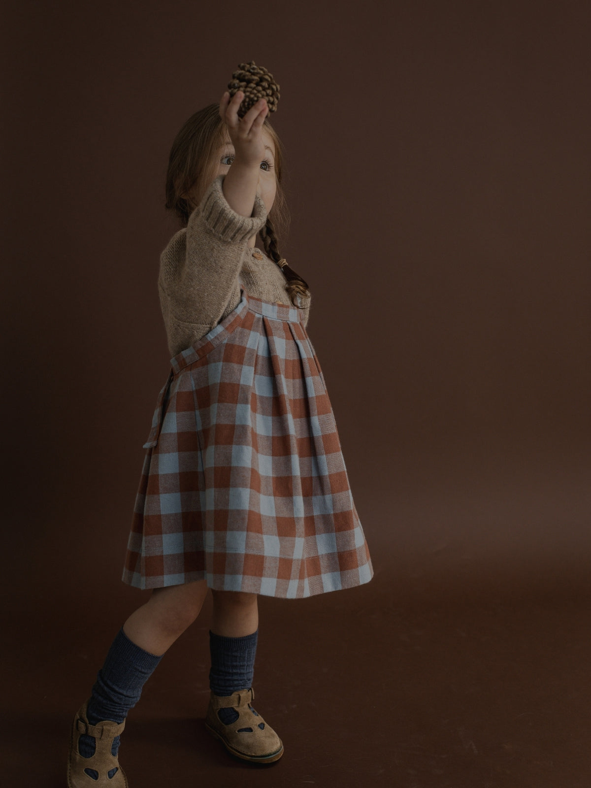 A young girl stands against a dark brown studio background wearing a brown and light blue plaid pinafore dress over a beige knit sweater, with dark blue socks and tan shoes.
