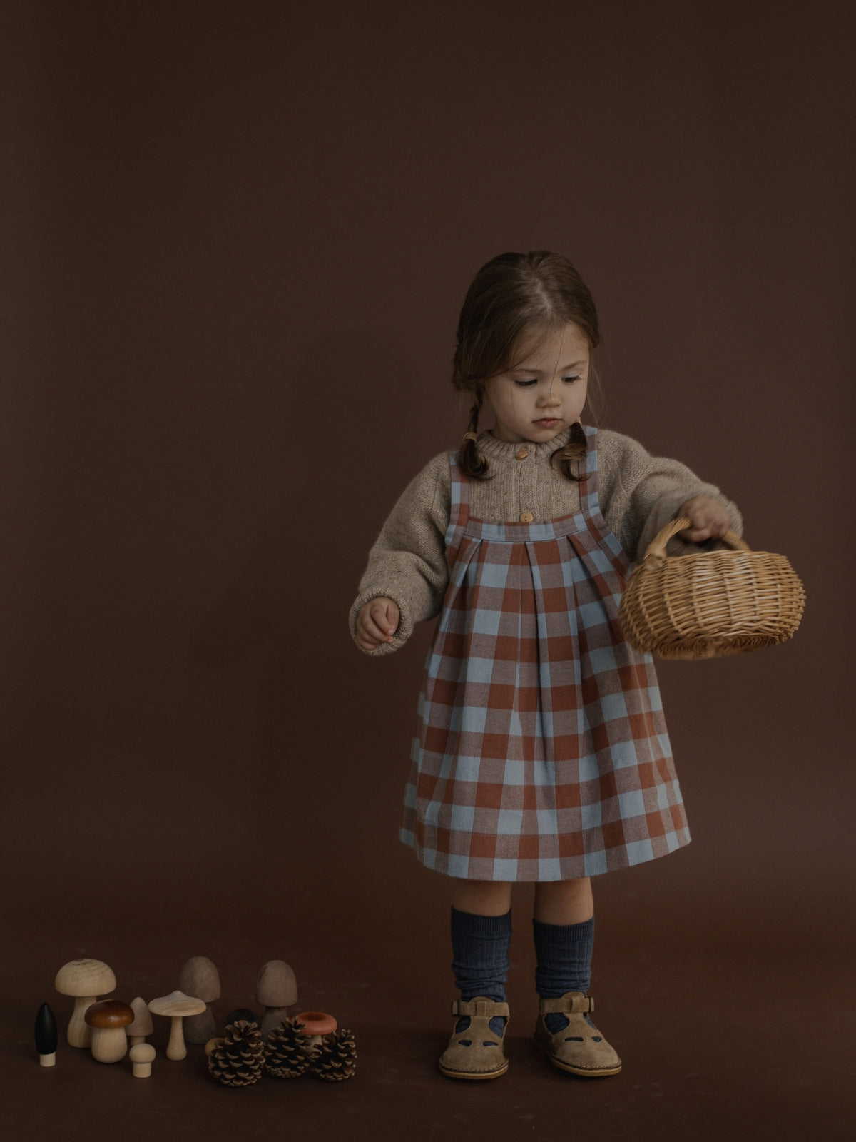 A young child stands against a dark brown studio backdrop wearing a brown and light blue plaid pinafore dress over a beige knit sweater, holding a small woven basket. A few toy mushrooms and pinecones are arranged on the floor nearby.