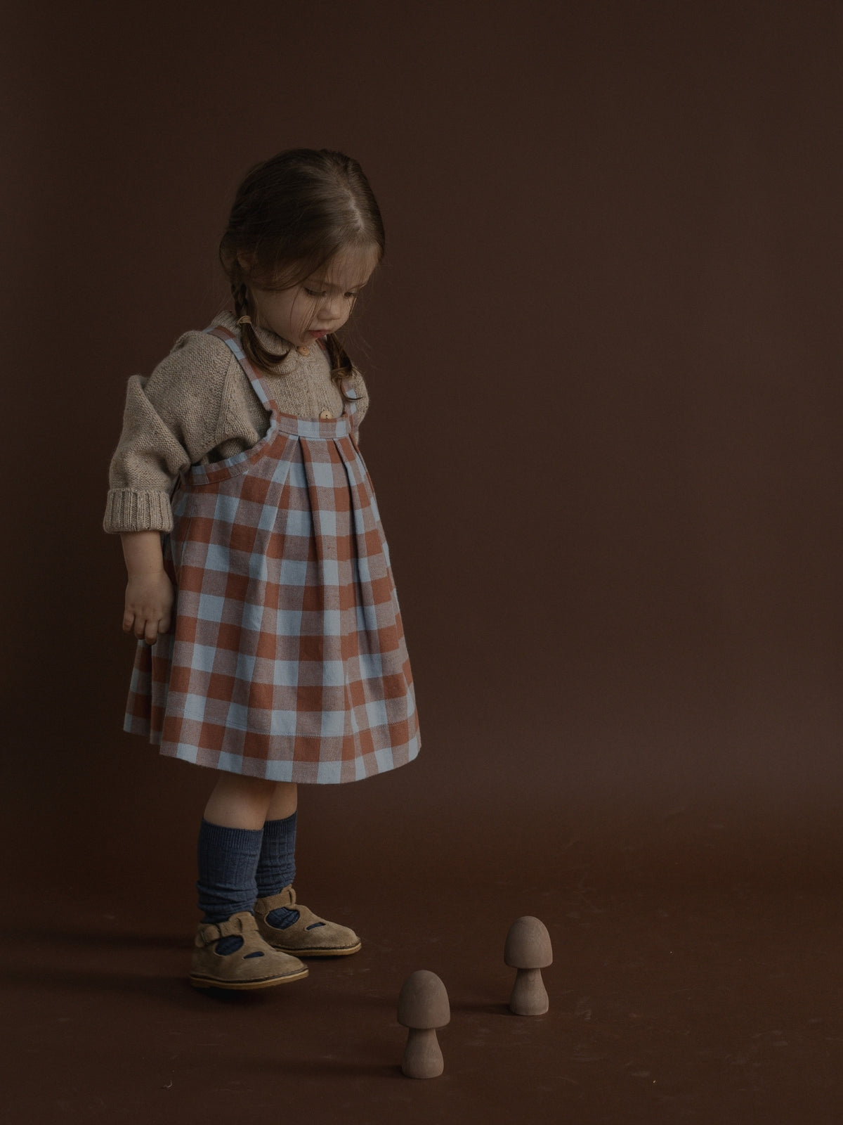 A young girl stands on a dark brown studio floor wearing a brown and light blue gingham pinafore dress over a beige knit sweater, with blue socks and tan shoes. Two small mushroom props sit on the floor in front of her against a solid dark brown backdrop.