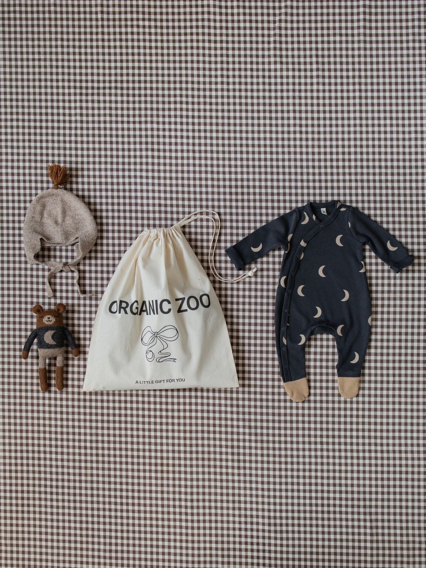 Baby outfit with navy blue romper, hat, and toys on a checkered background with Organic Zoo drawstring bag.