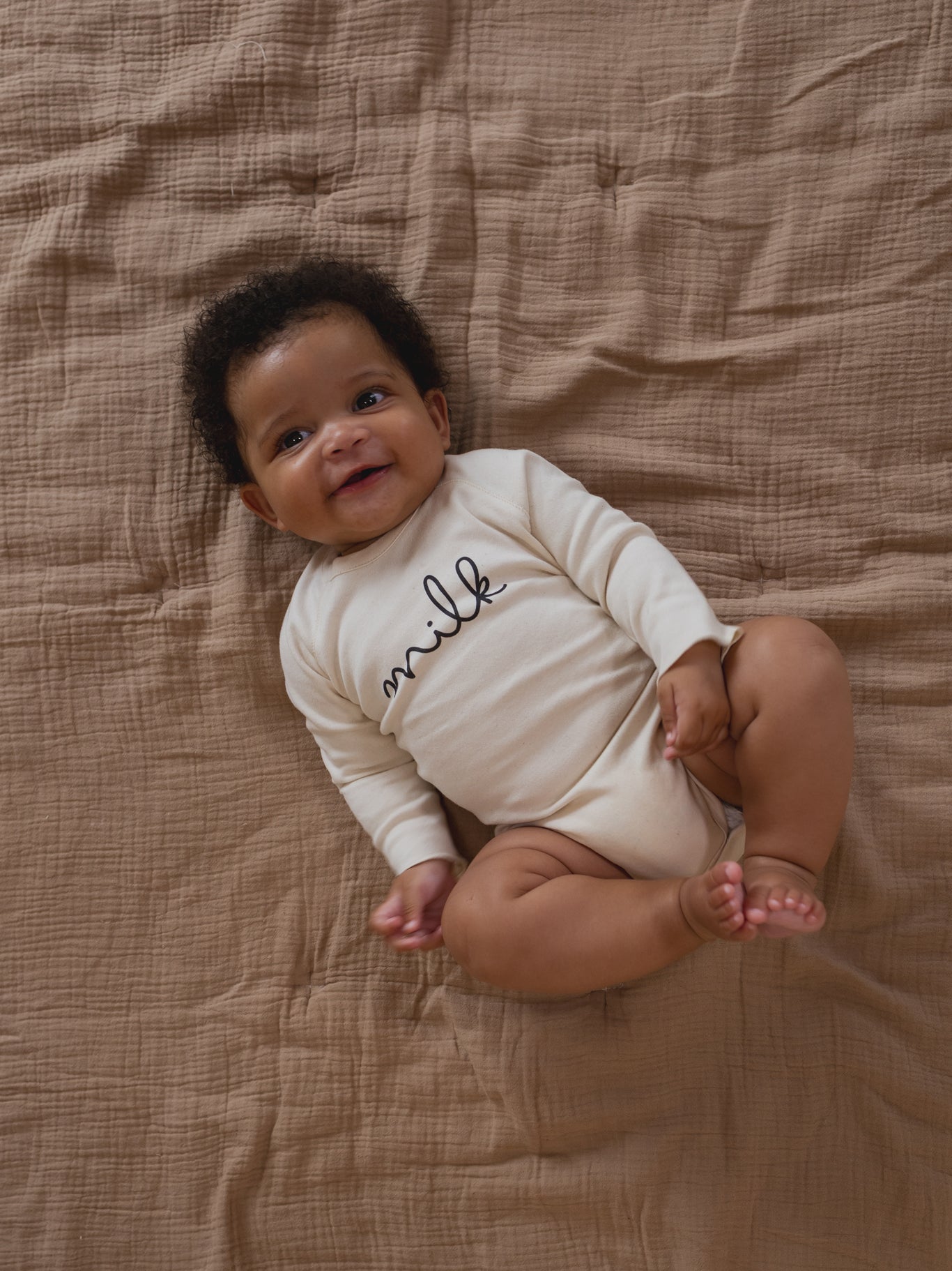 baby wearing cream undyed cotton bodysuit with milk text, lying on muslin blanket