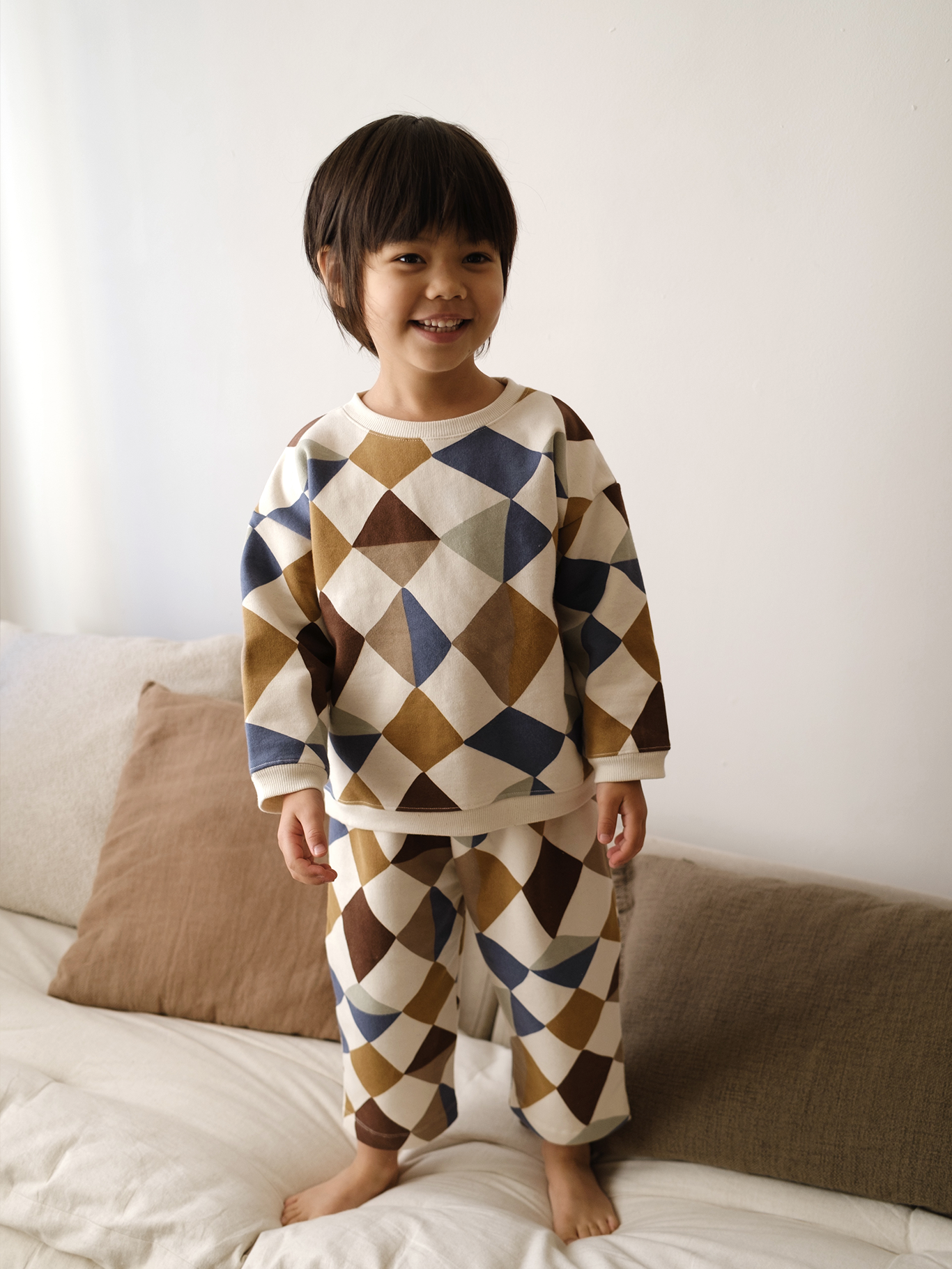 toddler in matching cream sweatshirt & pants with multicolour geometric patchwork print, standing on bed