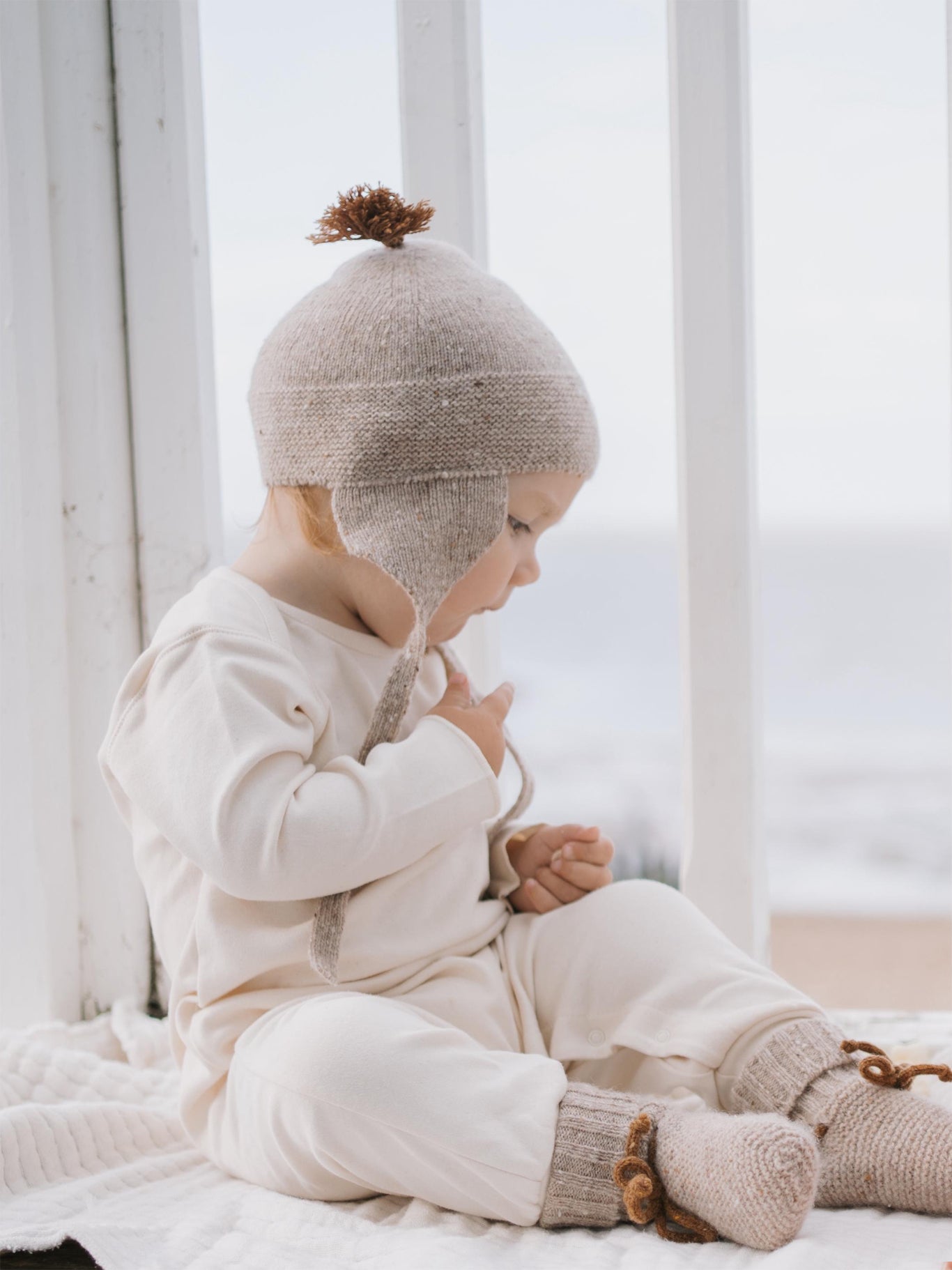 baby in wool booties in oatmeal with brown ribbons, with matching tassel hat and cream playsuit