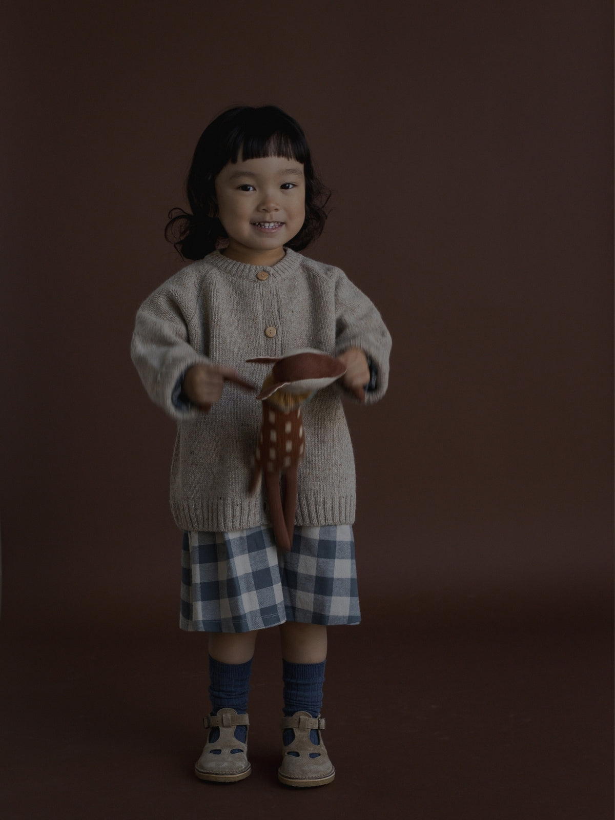 A young child stands against a dark brown studio background wearing a beige knit cardigan, a blue and white plaid skirt, blue socks, and beige shoes, while holding a brown and white patterned fabric accessory.