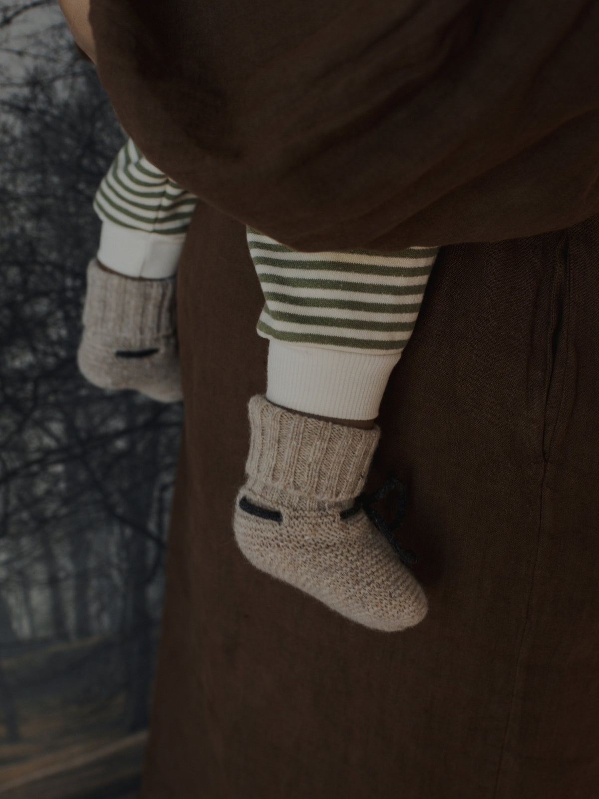 Close-up of a baby's legs dangling from a brown carrier or blanket, wearing striped green and white socks with beige knit booties.