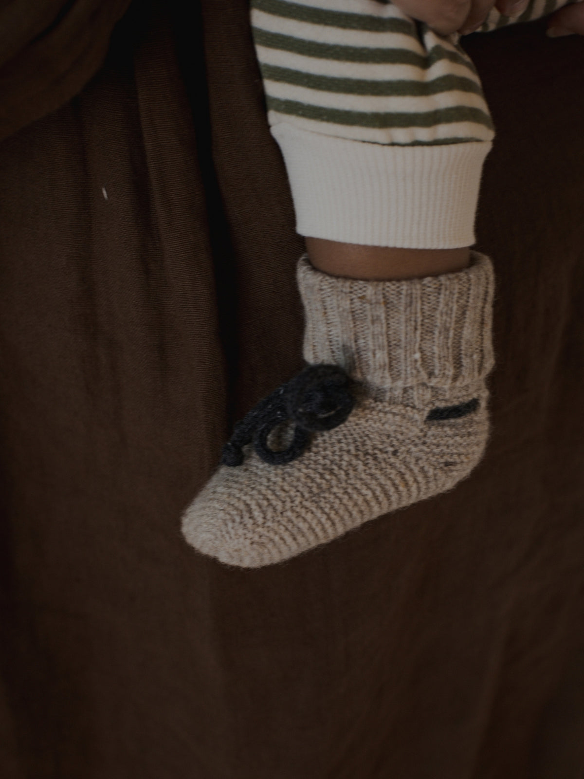 A close-up of a baby's foot wearing a beige knit bootie with a dark gray bow, held against a brown fabric background.