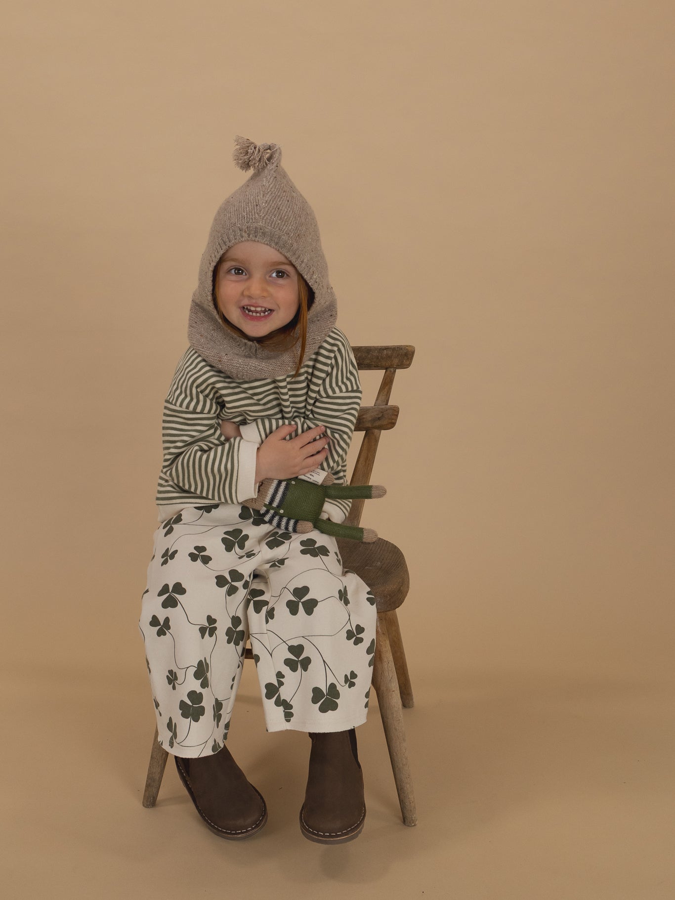 toddler in oatmeal wool knitted balaclava, green striped sweatshirt and clover pants, hugging toy