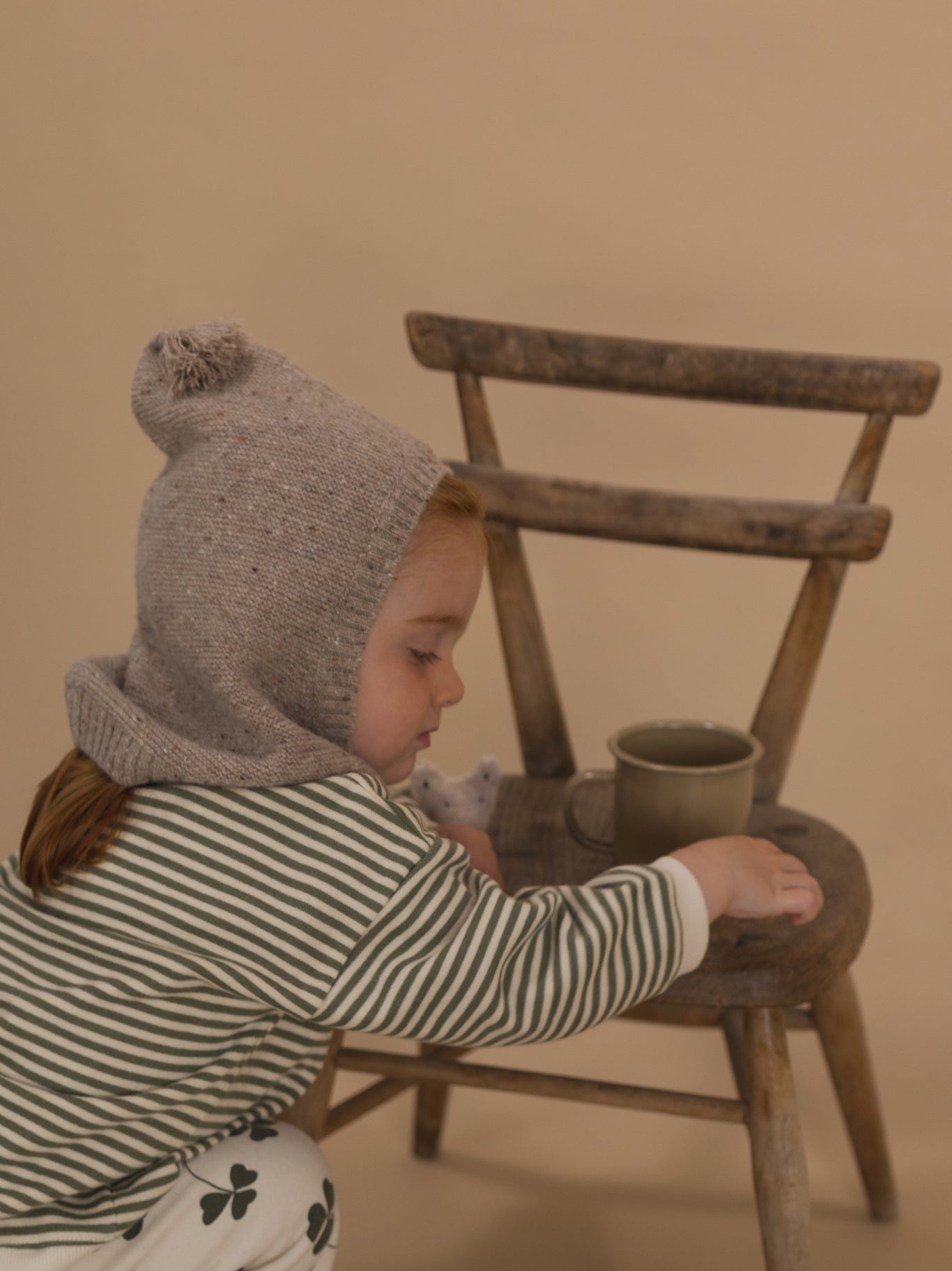 toddler in oatmeal wool knitted balaclava, striped sweatshirt and clover pants, playing on chair