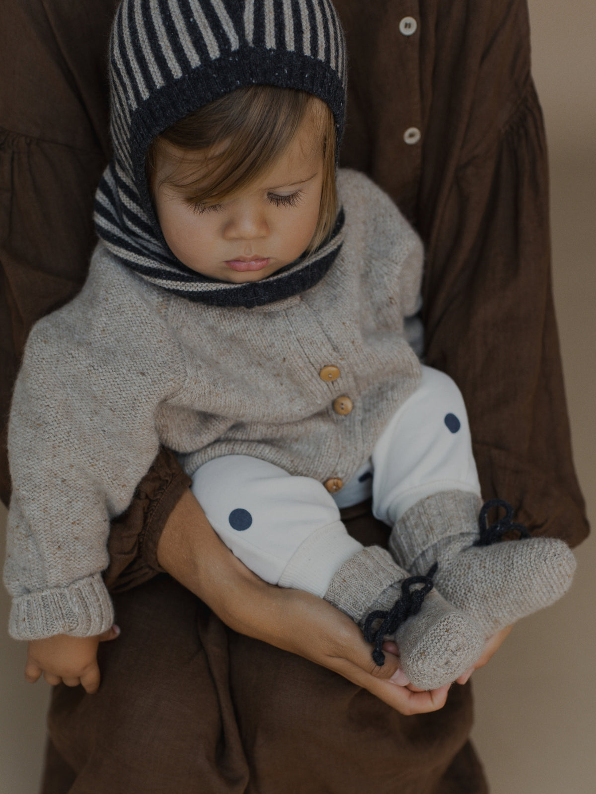 A baby wearing a cosy knit outfit including a striped bonnet, grey cardigan, white pants, and grey booties, held by an adult in a brown shirt against a neutral background.