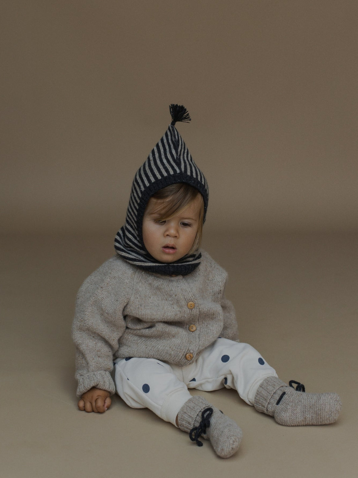 A baby sitting on a warm beige studio background, wearing a gray knit cardigan, white pants with blue polka dots, gray knit socks, and a navy-and-white striped bonnet with a black pom-pom.
