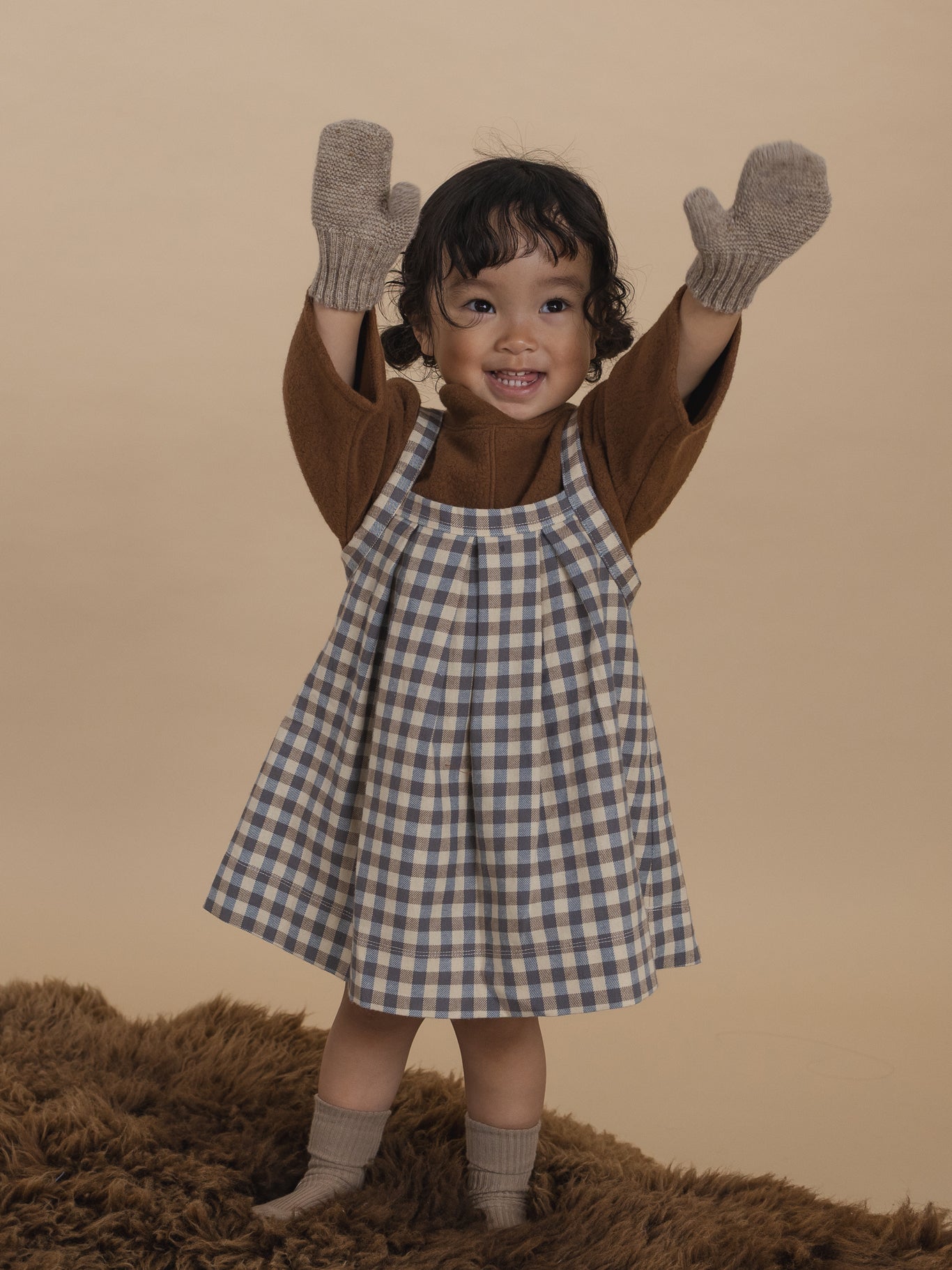 toddler in oatmeal mittens with ribbed cuffs, brown jumper and gingham skirt standing with hands up