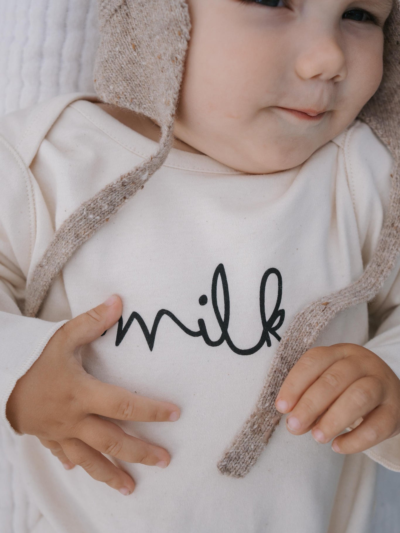 close up of a baby in cream undyed cotton playsuit with milk text, and oatmeal wool hat