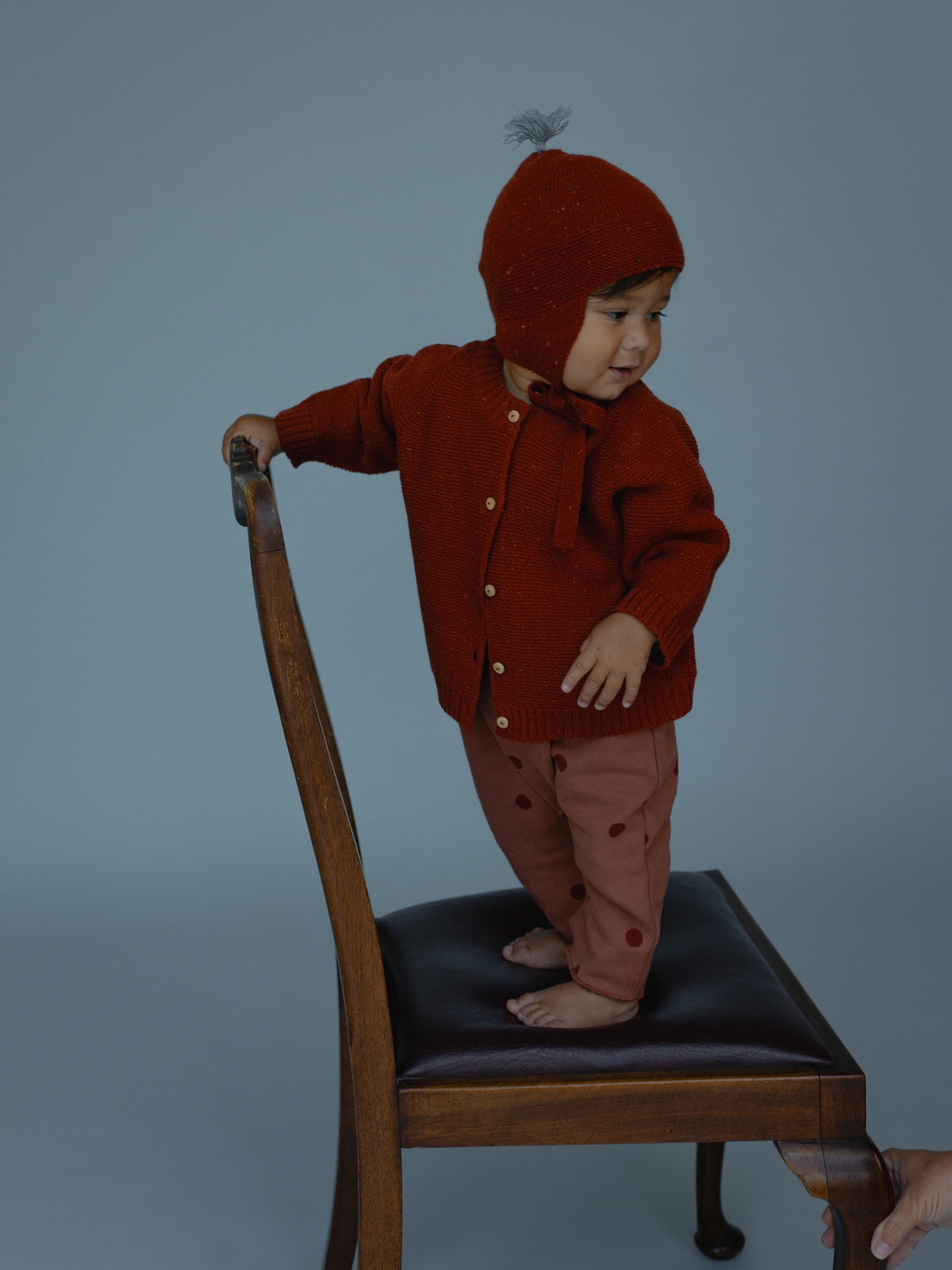 A toddler stands on a wooden chair wearing a rust-red knit cardigan and matching bonnet with a pom-pom, paired with dusty pink pants with red dots, against a solid blue-gray studio background.