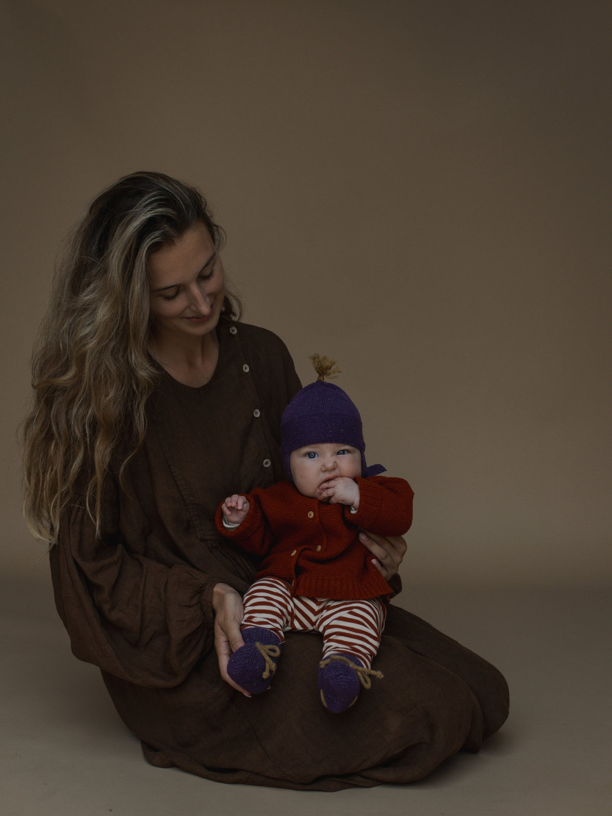 A woman in a brown long-sleeve top sits on the floor holding a baby dressed in a red knit cardigan, purple hat, and red-and-white striped pants against a plain brown studio background.