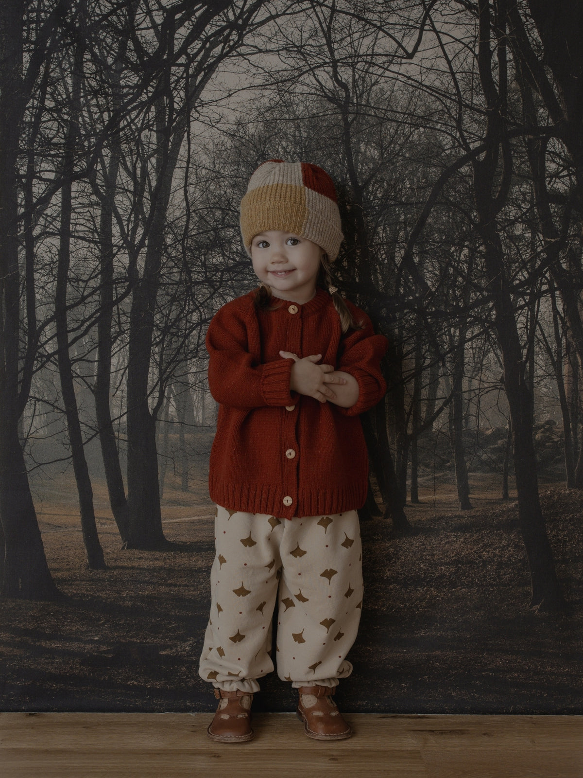 A toddler stands in front of a forest backdrop wearing a rust red knit cardigan, cream knit pants with small brown animal prints, and a beige and red knit beanie.