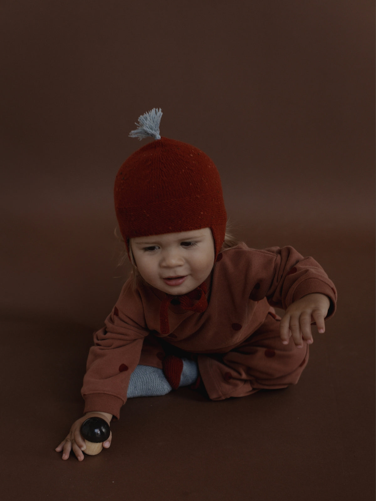 A baby wearing a rust-red knit bonnet with a white tassel and a matching rust-brown romper, crawling on a dark brown studio background while holding a wooden toy.