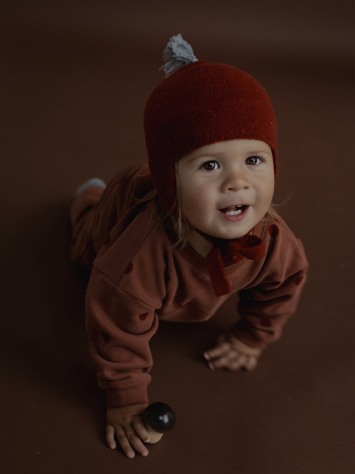A baby crawling on a dark brown studio floor, wearing a rust-brown long-sleeve romper and a matching knit bonnet with a small gray pom-pom.