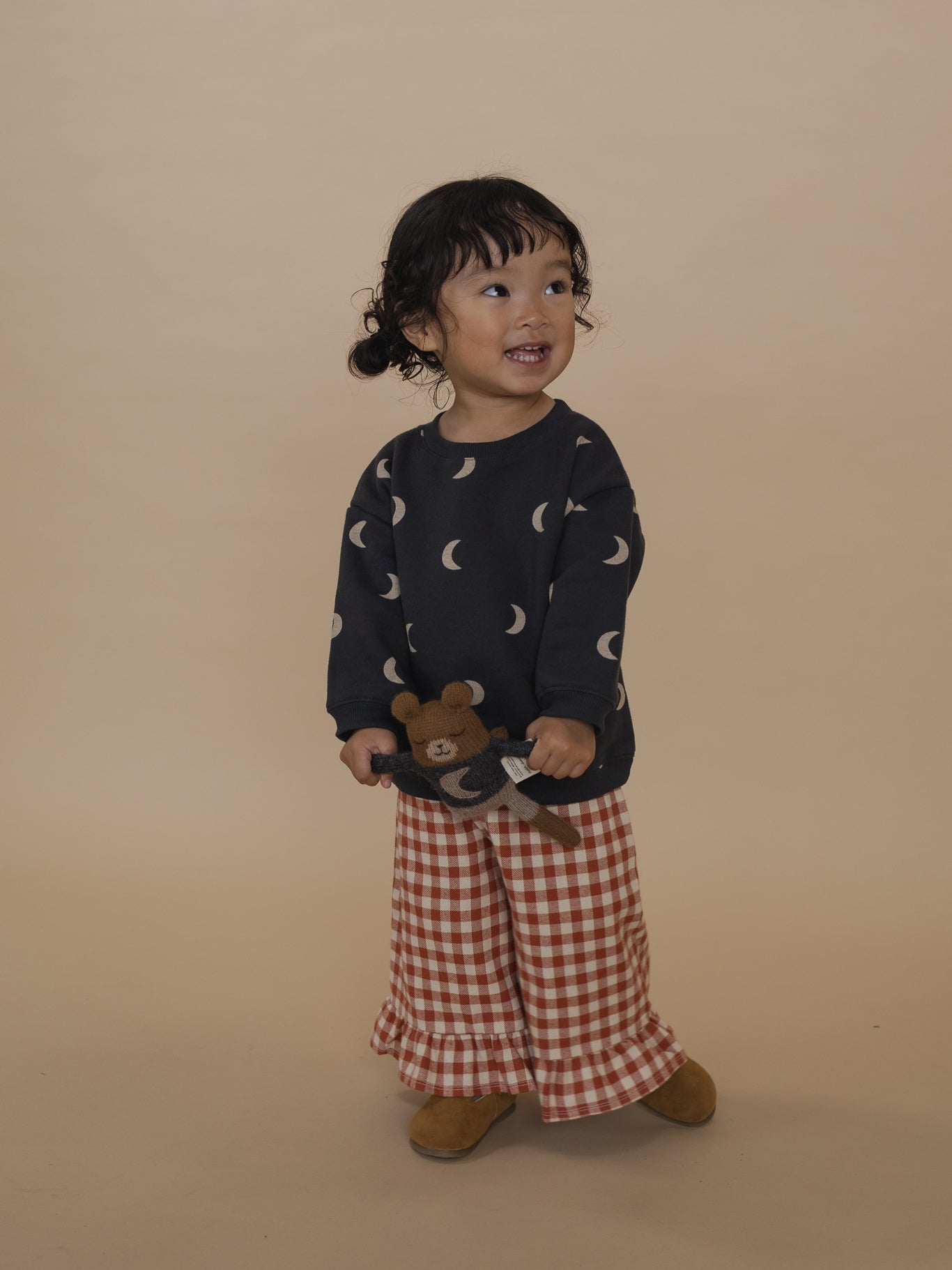 toddler in red gingham culottes with frill hems and black sweatshirt with moon print, holding a plushie