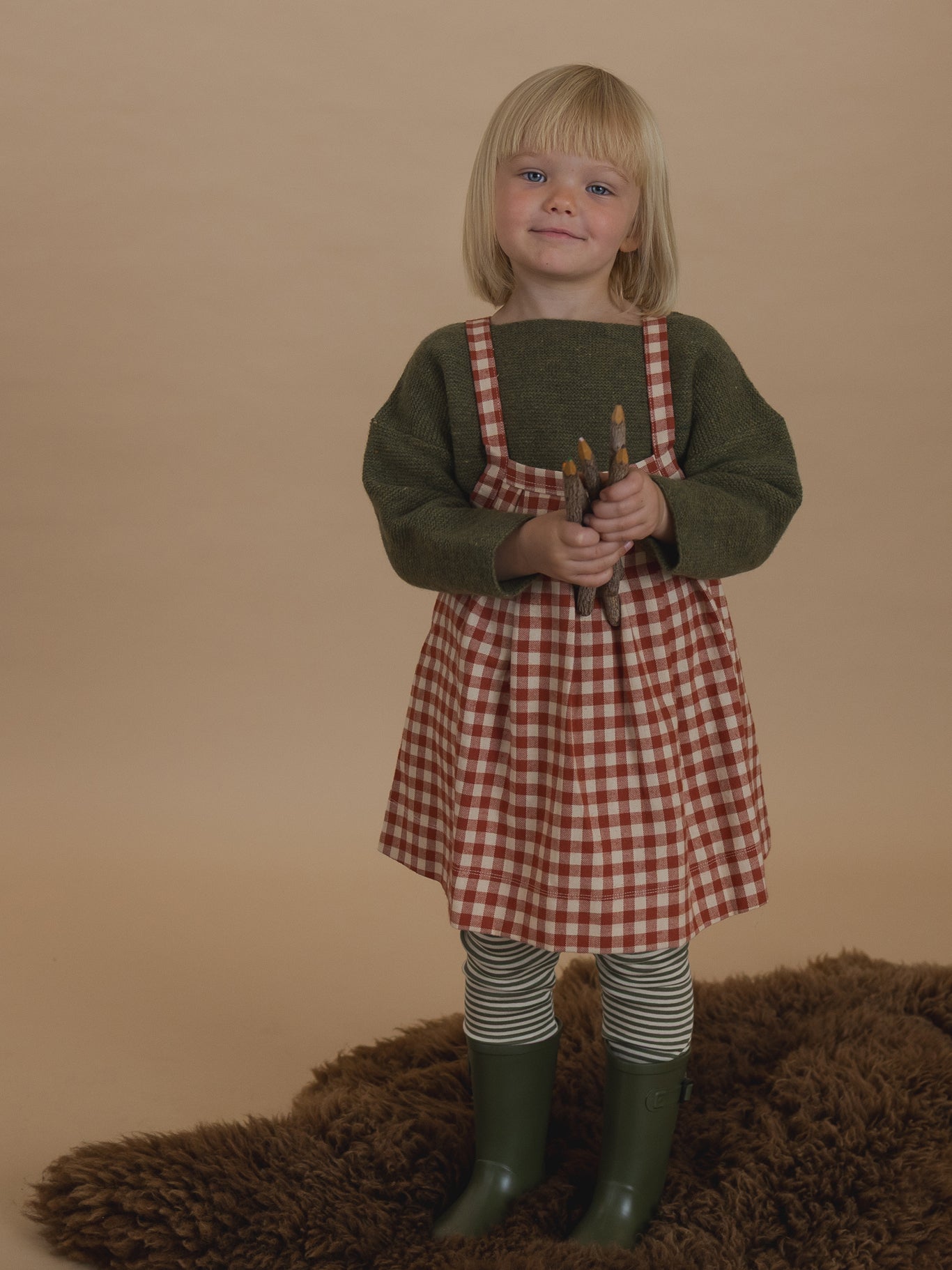 toddler in paprika red gingham pinafore skirt with green wool jumper, holding a bunch of pencils