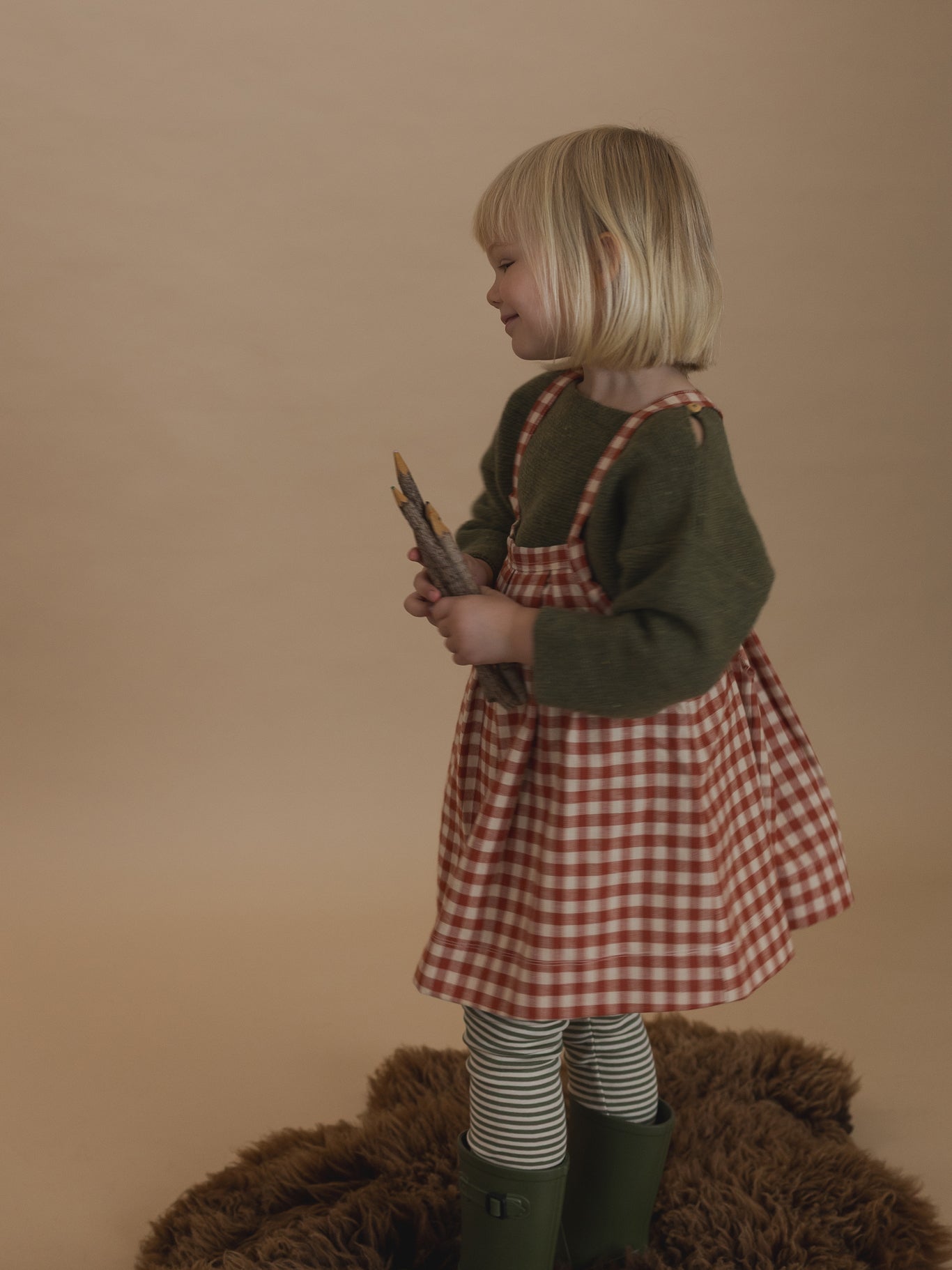 toddler in red gingham tribe skirt with wool jumper and striped sweatpants, holding coloured pencils