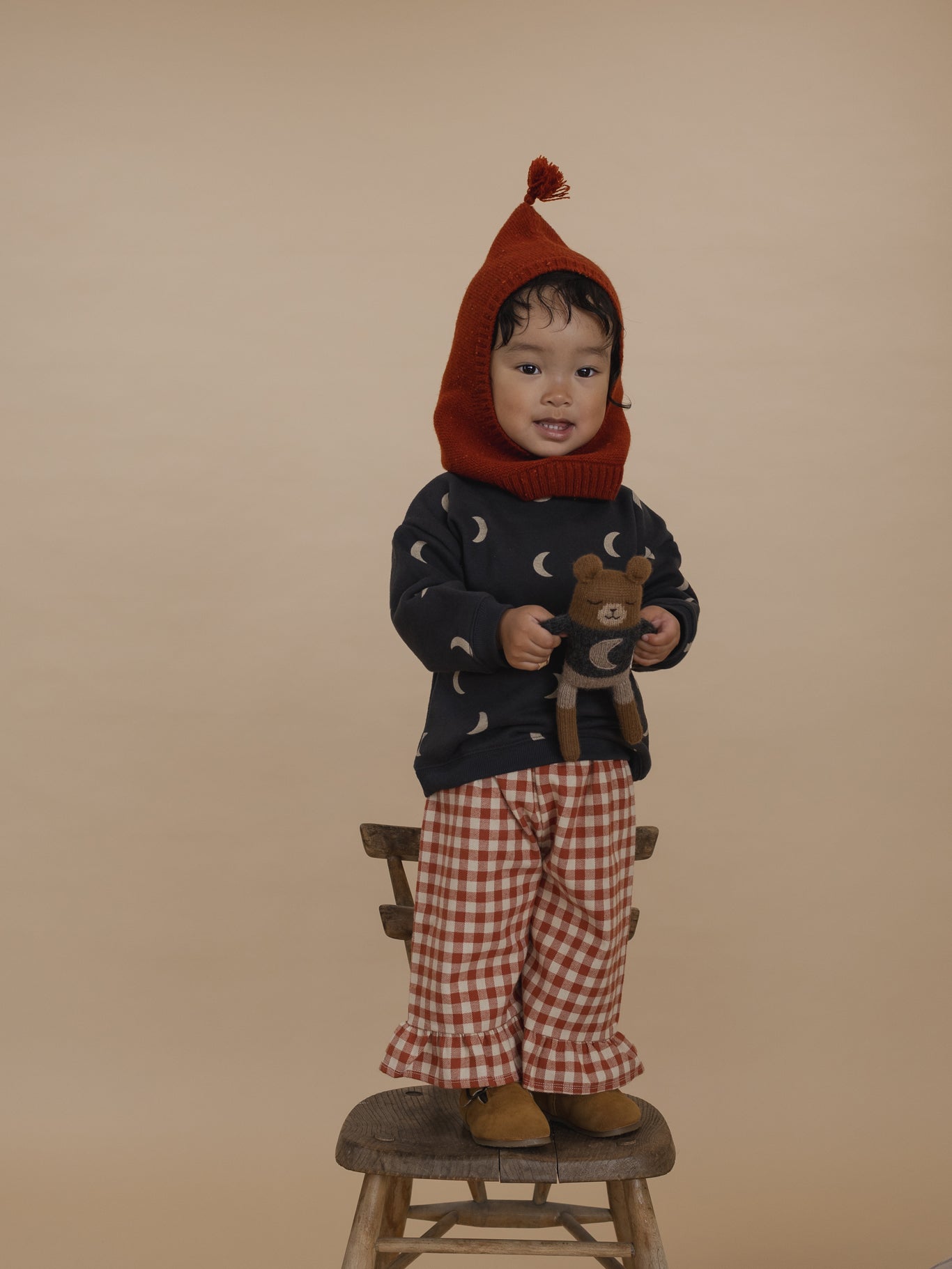 toddler in red wool balaclava with tassel, moon sweatshirt, gingham culottes, standing on chair with toy