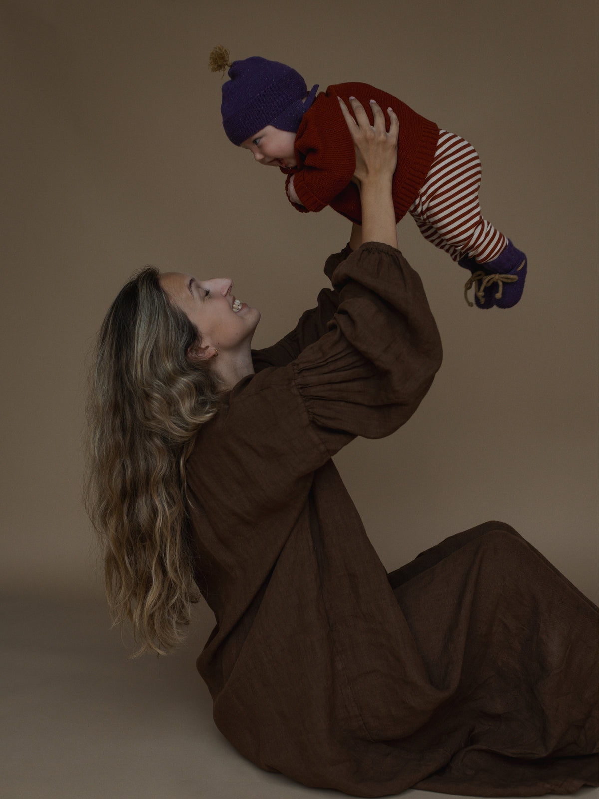 A woman in a brown long-sleeved dress kneels on a beige studio floor, reaching up to hold a baby wearing a purple hat, red top, and red-and-white striped pants.
