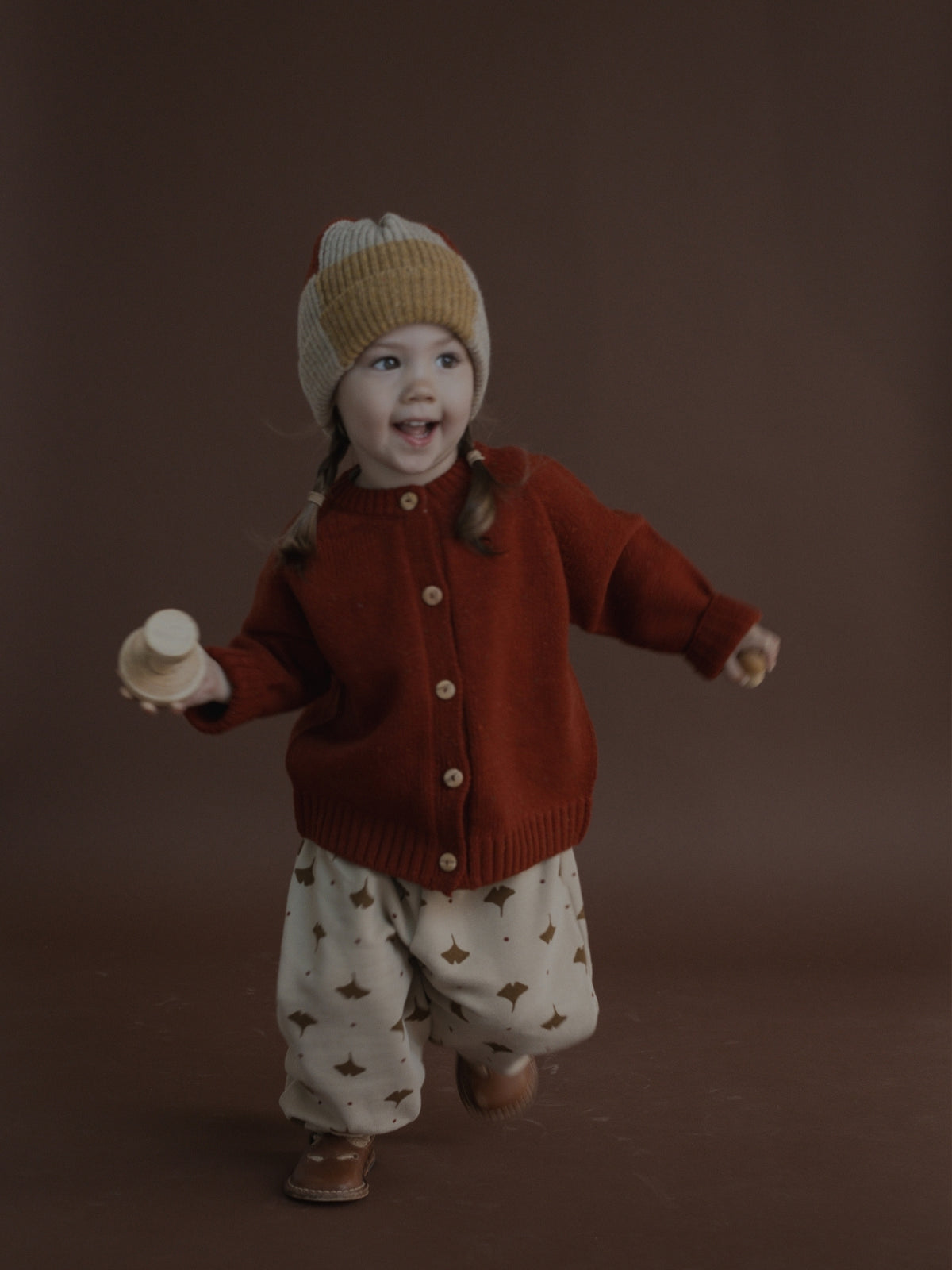 A toddler wearing a rust-red button-up cardigan, cream knit pants with brown star patterns, and a beige knit beanie, smiling and holding a small wooden toy against a solid dark brown studio background.
