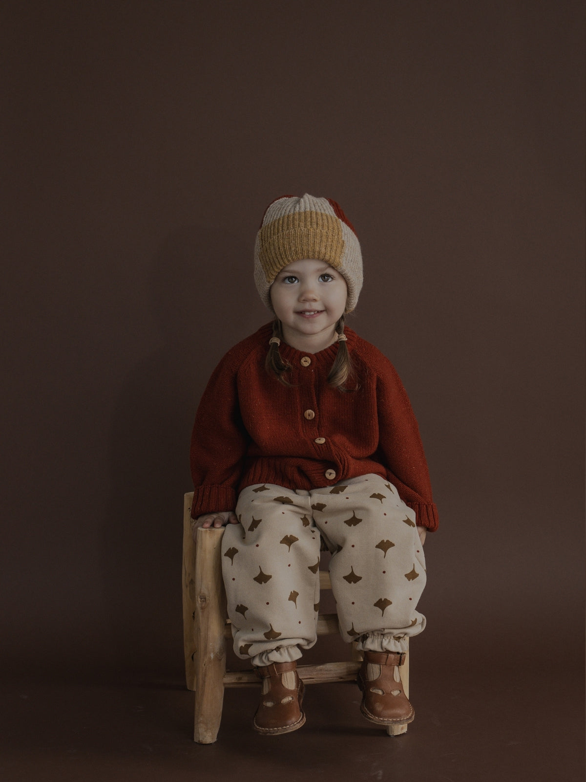 A young child sitting on a small wooden stool, wearing a rust red cardigan, cream pants with brown geometric patterns, and a beige knit beanie with a red stripe, against a solid dark brown studio background.