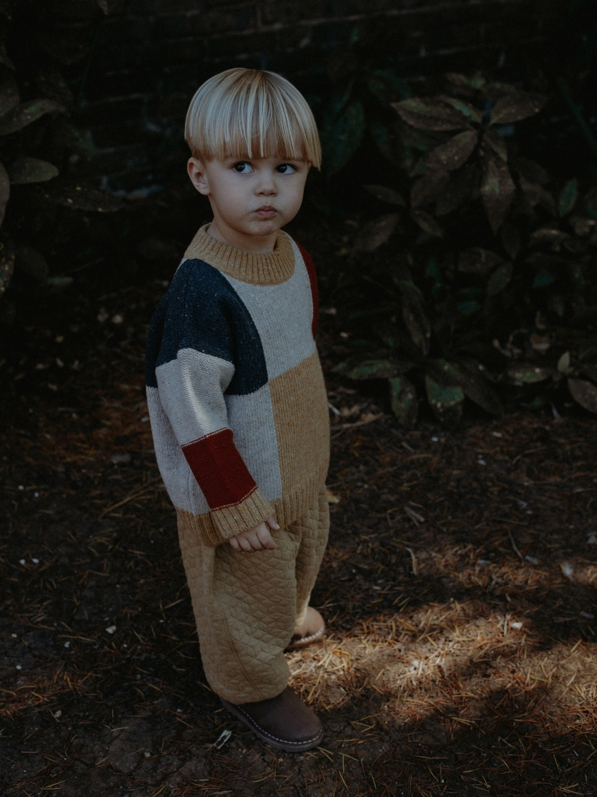 A young child wearing a color-block knit jumpsuit in beige, cream, navy, and red, standing outdoors on a dark forest floor with pine needles and foliage.