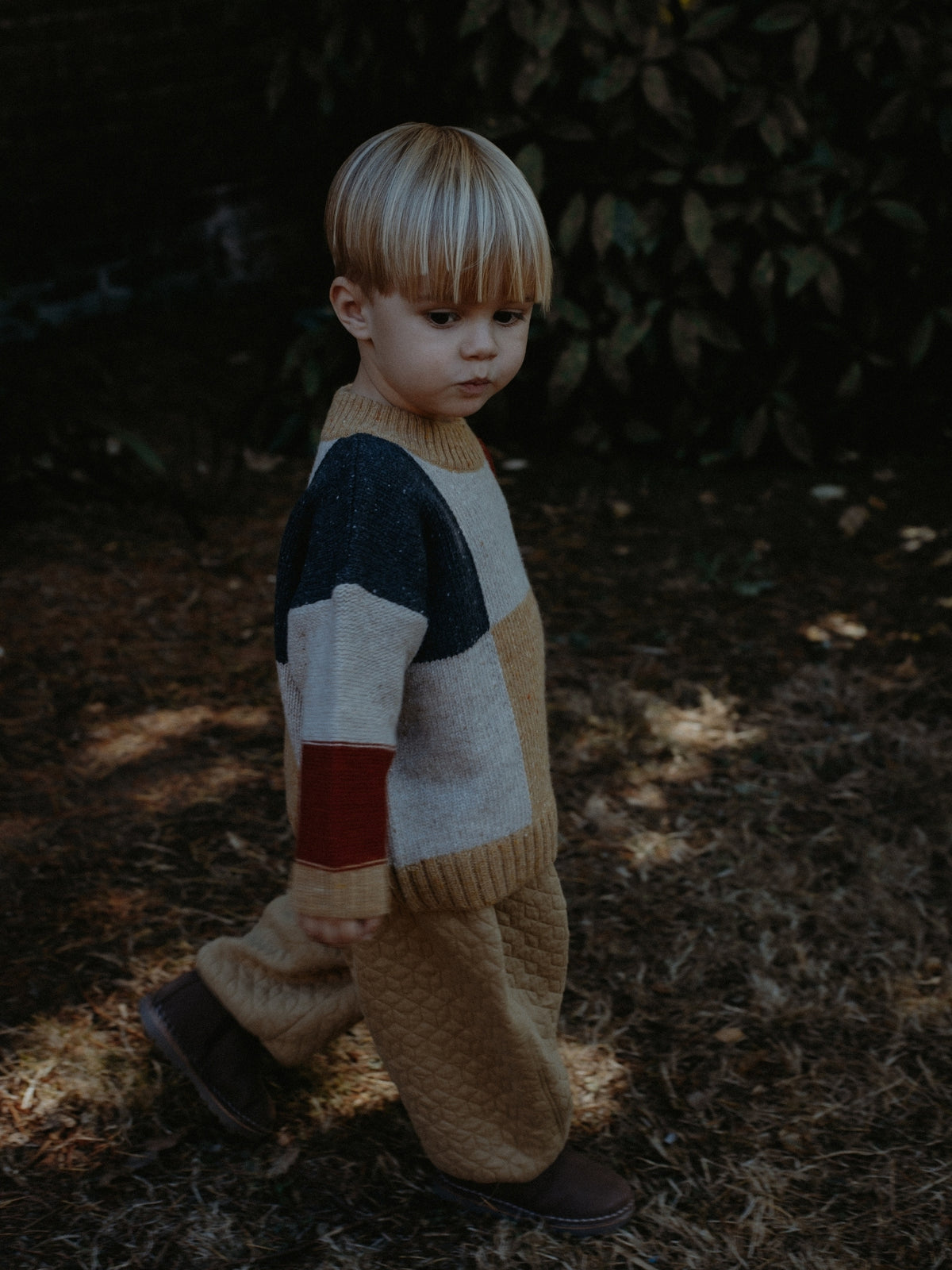 A young child wearing a color-block knit sweater and matching knit pants stands outdoors on a wooded path. The outfit features large blocks of cream, tan, dark navy, and a muted red on the sleeves, paired with matching tan knit pants.