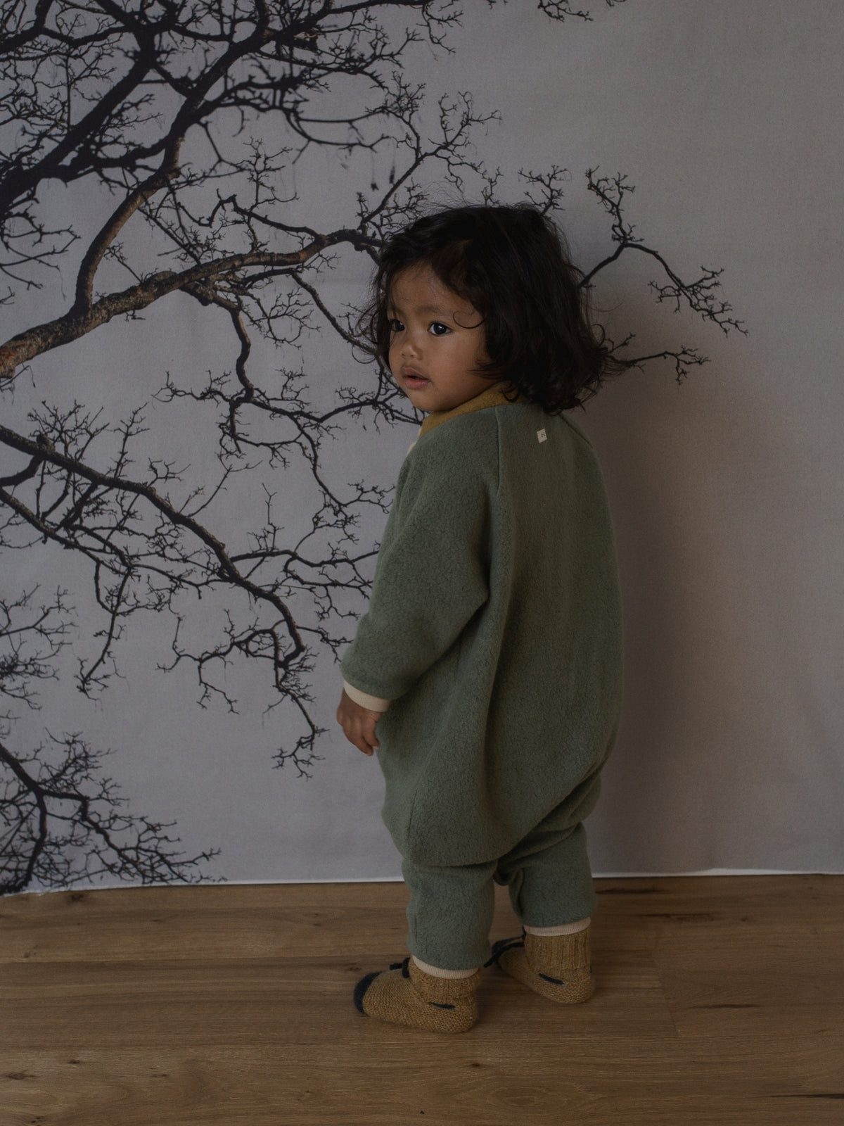 A toddler stands with their back to the camera wearing a sage green knit romper and matching pants, standing on a wooden floor in front of a wall with a black tree branch print.