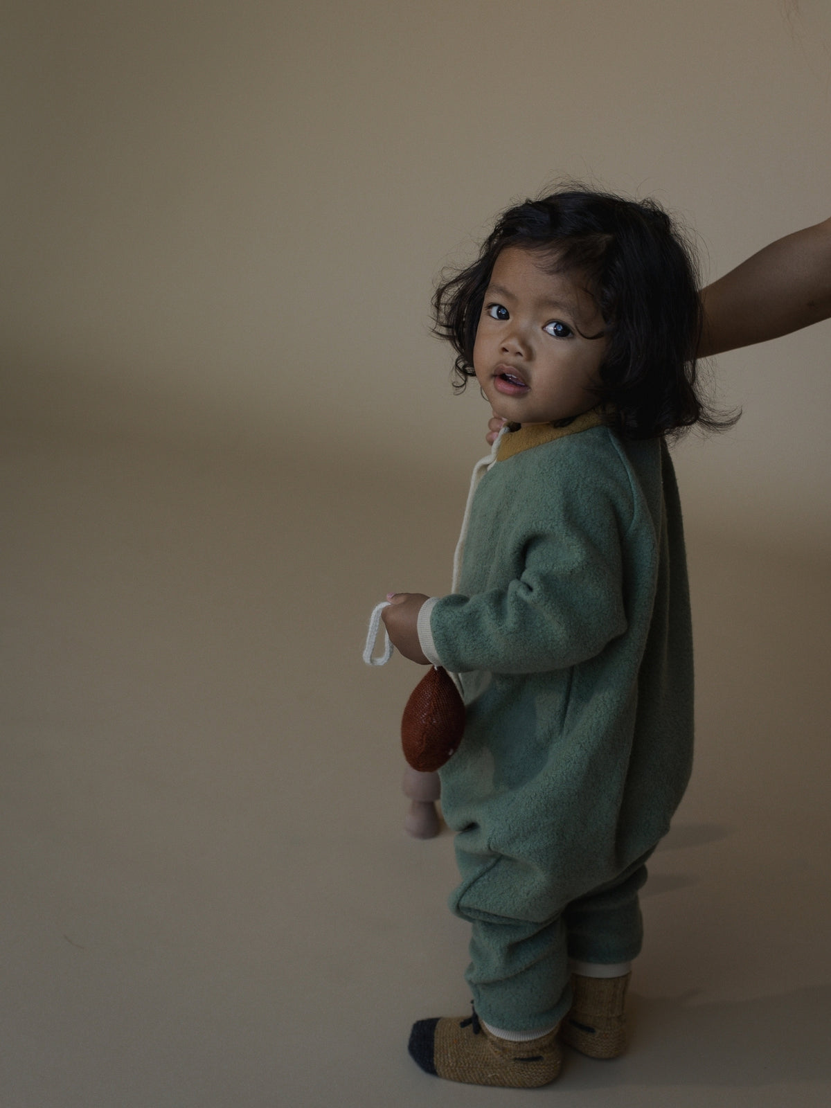 A toddler stands against a plain beige studio background wearing a sage green fleece one-piece outfit with white trim and tan socks. A hand is visible behind the child, and the child holds a small brown object.