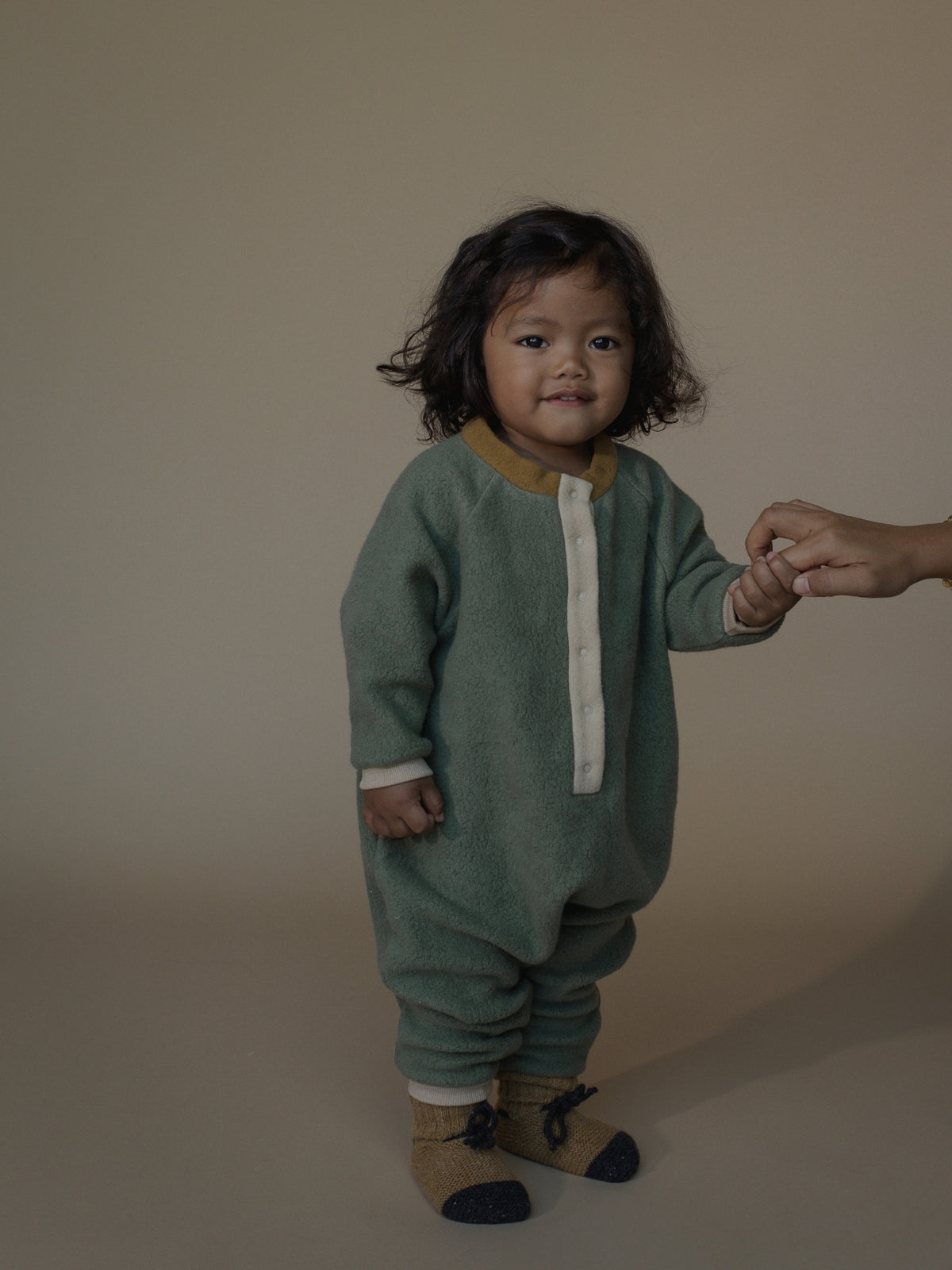 A toddler wearing a sage green knit romper with a mustard yellow collar and white front placket, standing and holding an adult's hand against a plain beige studio backdrop.