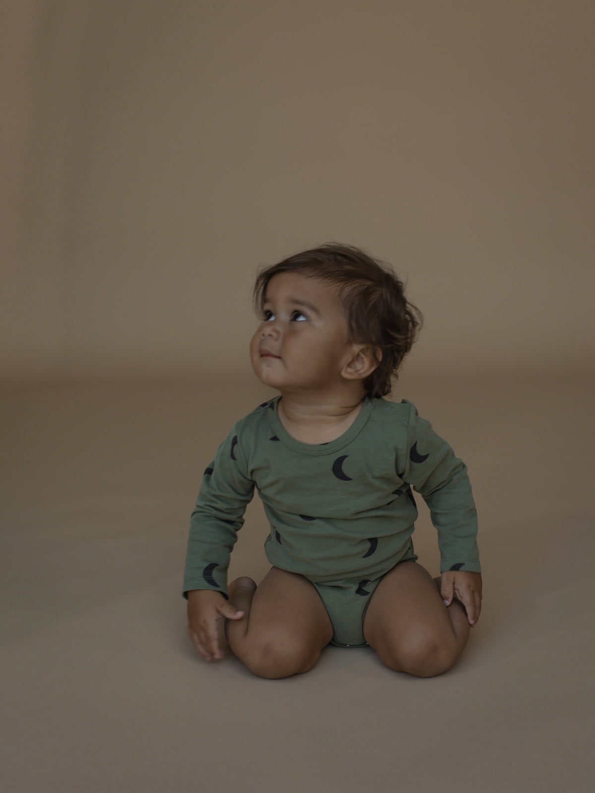 A baby sitting cross-legged on a warm beige studio floor, wearing a sage green long-sleeve romper with a dark crescent moon pattern.