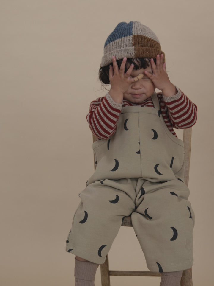 toddler in wool jumper with paprika red stripes, beige dungarees and patchwork beanie, playing peekaboo