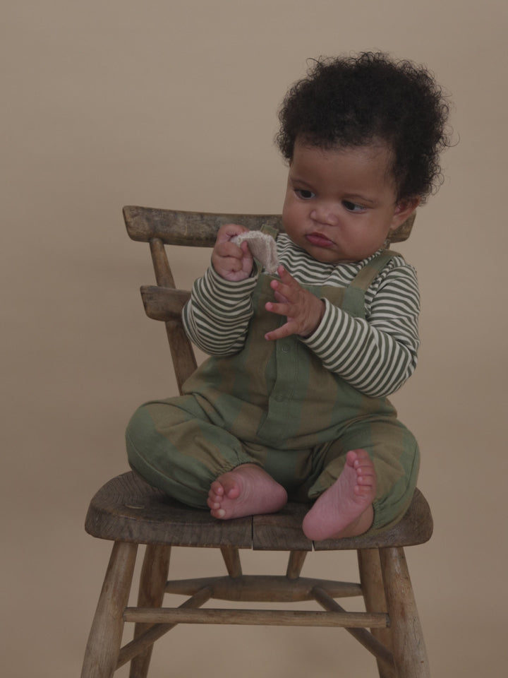 baby in green and cream gingham salopette and striped sweatshirt, playing with toy on a chair