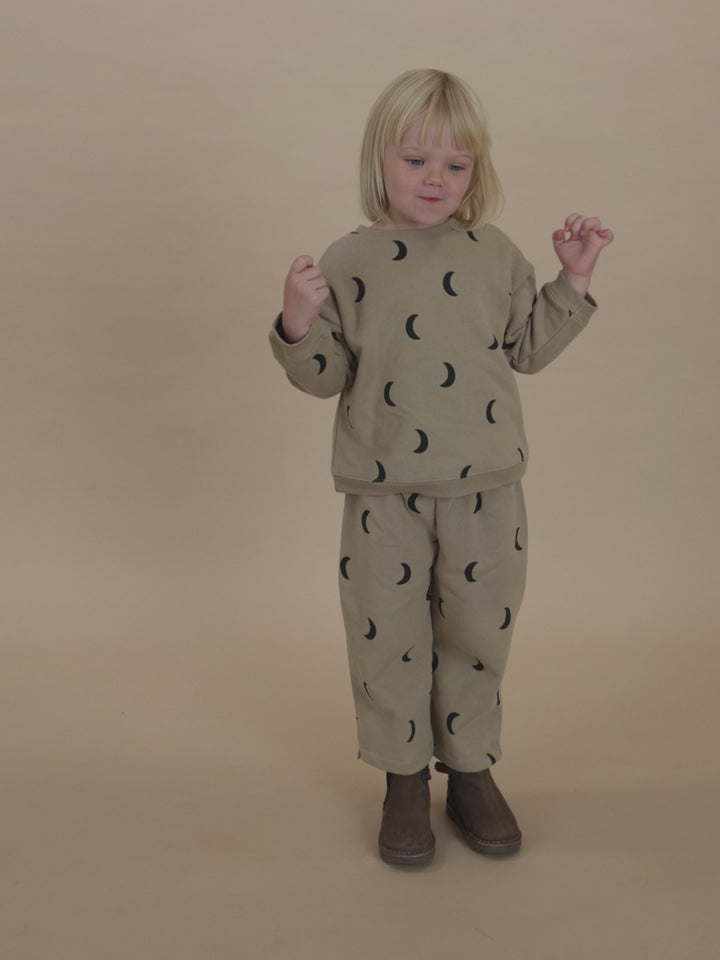 toddler in dusky beige sweatshirt with black moon print & matching pants, posing on one leg with arms out