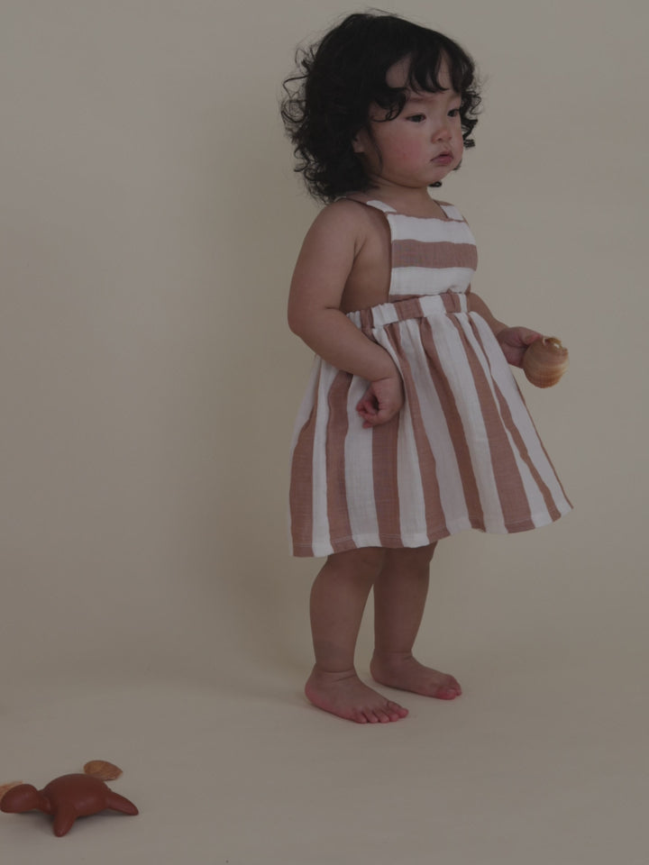 toddler in red striped apron skirt placing seashells in pink mesh basket, picking toy turtle