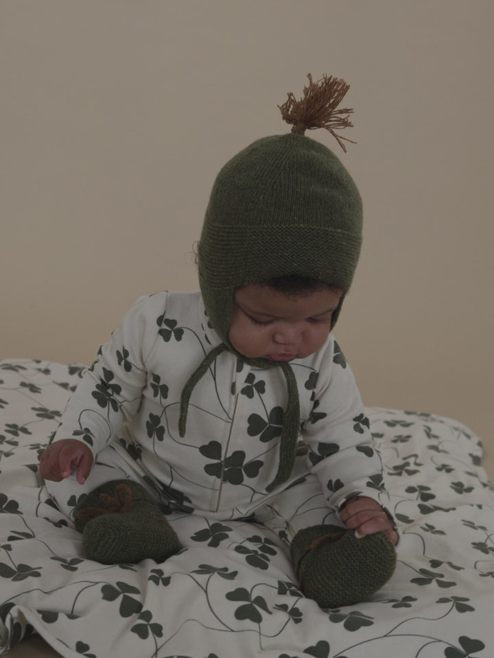 baby sitting on blanket with green clover print, in matching playsuit, green wool hat and booties