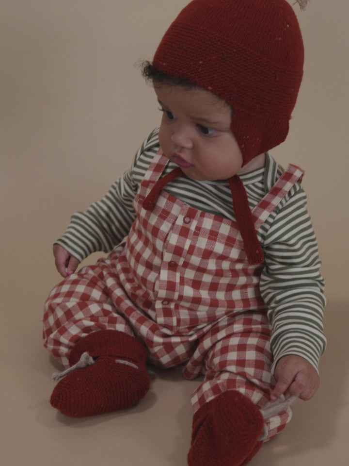 baby wearing red wool tassel hat with chin tie, matching booties, gingham dungarees and striped top