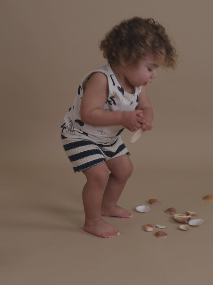toddler in white summer vest with ocean print & navy striped knitted shorts picking seashells