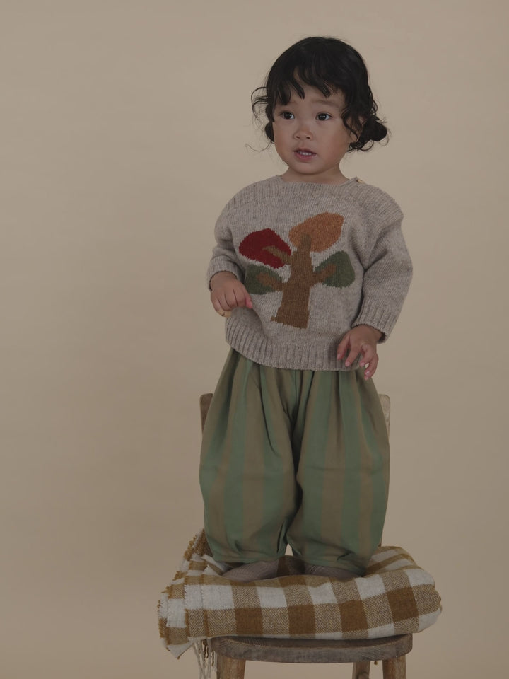 toddler in beige wool jumper with tree motif and striped balloon pants, standing on chair clapping
