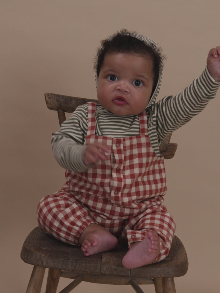 Baby sitting on wooden stool wearing checkered outfit and green striped bonnet, playing with hands