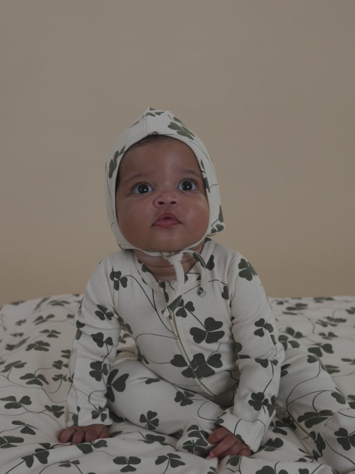baby in matching cream pixie bonnet and onesie with green clover print, sitting on matching blanket