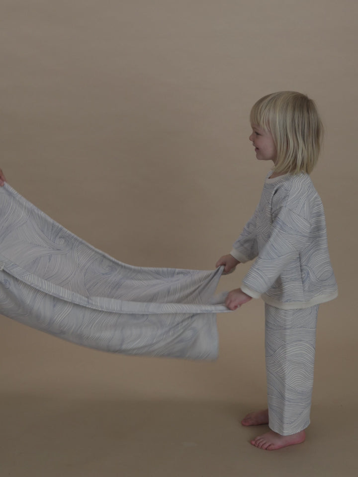 toddler folding a cream blanket with blue line-drawn ocean waves, in matching sweatshirt and pants