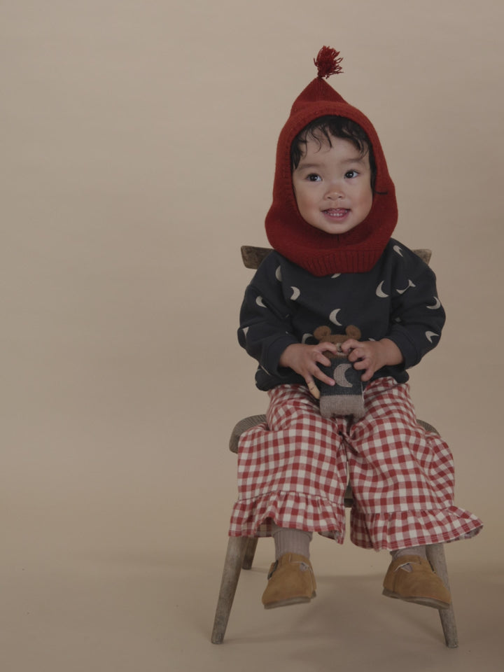 toddler in red wool balaclava, moon sweatshirt, gingham frill culottes, swinging legs