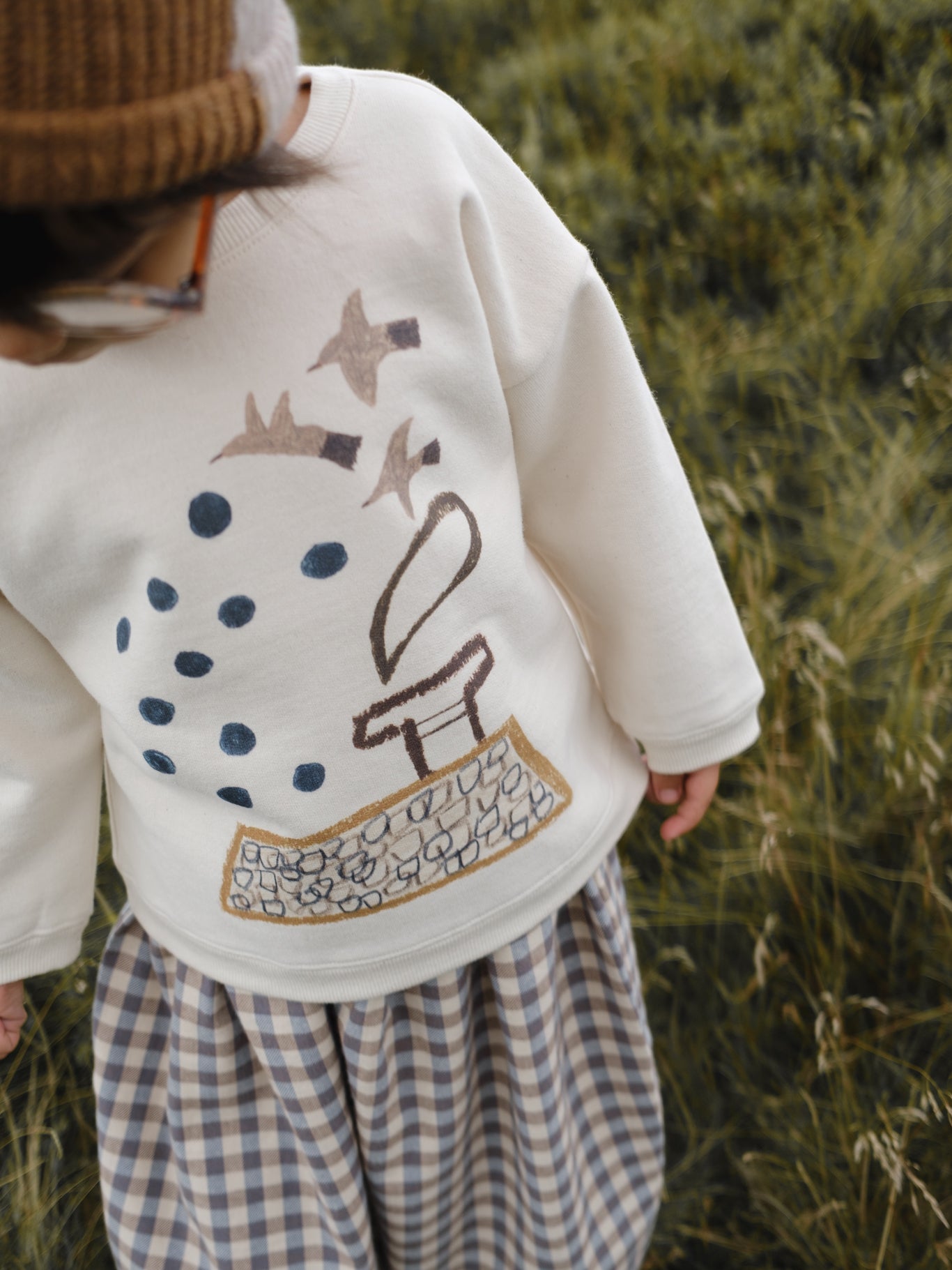 close up of toddler in cream sweatshirt with birds and blue dots, gingham balloon pants in grassy meadow